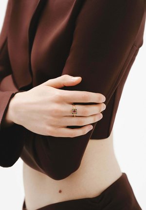 Anillo de oro con una piedra central roja rectangular, flanqueada por piedras verdes más pequeñas. El modelo lleva una blusa ajustada de color marrón oscuro. La piel es de tono claro.