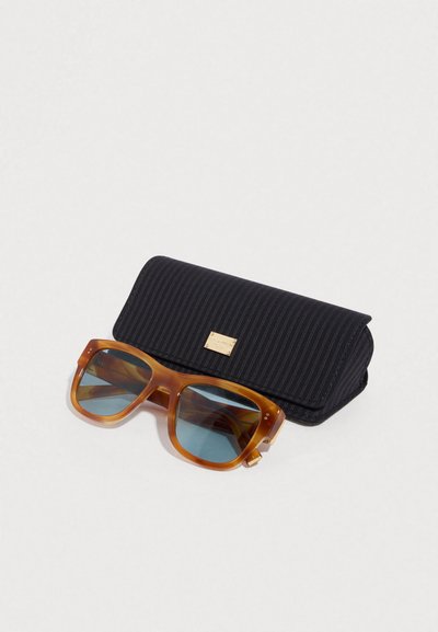 Γυαλιά ηλίου με σκελετό σε χρώμα χελώνας και μπλε φακούς, τοποθετημένα σε μια μαύρη θήκη με ρίγες και χρυσή διακόσμηση. Η θήκη διαθέτει κλείσιμο με πτερύγιο.