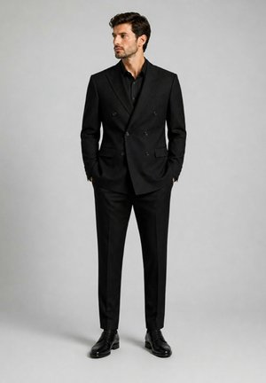 Homme en costume noir croisé avec une chemise noire et des chaussures noires brillantes, debout devant un fond gris uni.