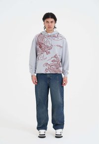 Felpa con cappuccio grigia chiara decorata con dettagli grafici di un drago rosso e nuvole, presenta cappuccio con cordini e polsini a coste, abbinata a jeans a gamba larga blu.