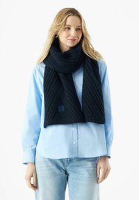 Écharpe en tricot marine avec un motif en rib texturé, arborant un petit accent de logo bleu, portée avec une chemise bleue clair à boutons.