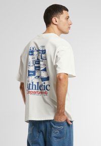Tricou alb din bumbac, cu un grafic al cutiilor de pantofi stivuite în nuanța albastră pe spate, cu textul "Departamentul Sportiv" dedesubt. Design ajustat.