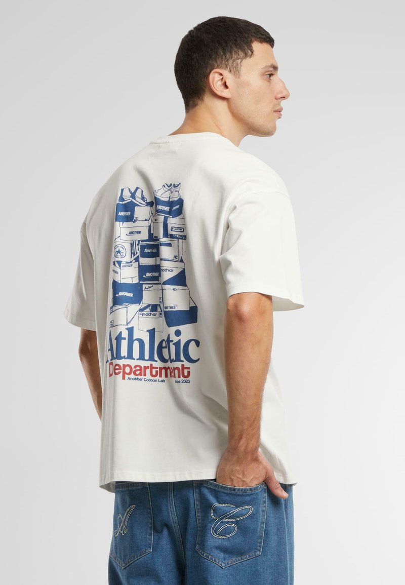 Tricou alb din bumbac, cu un grafic al cutiilor de pantofi stivuite în nuanța albastră pe spate, cu textul "Departamentul Sportiv" dedesubt. Design ajustat.