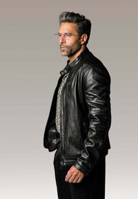 Veste de moto en cuir noir avec des épaules texturées, des détails en zip et un design ajusté. Portée par-dessus une chemise à motifs. Des lunettes complètent le look.