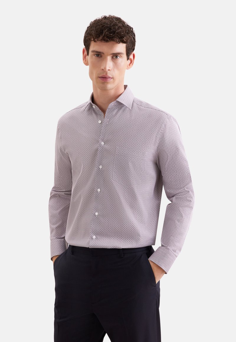 Homme aux cheveux bouclés et foncés, portant une chemise claire à motifs à manches longues boutonnée et un pantalon noir, debout les mains dans les poches.
