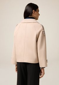 Lichtbeige wollen jack met een gestructureerde kraag, gegelateerde schouderaccenten en korte mouwen. Zachte textuur en een comfortabele pasvorm.