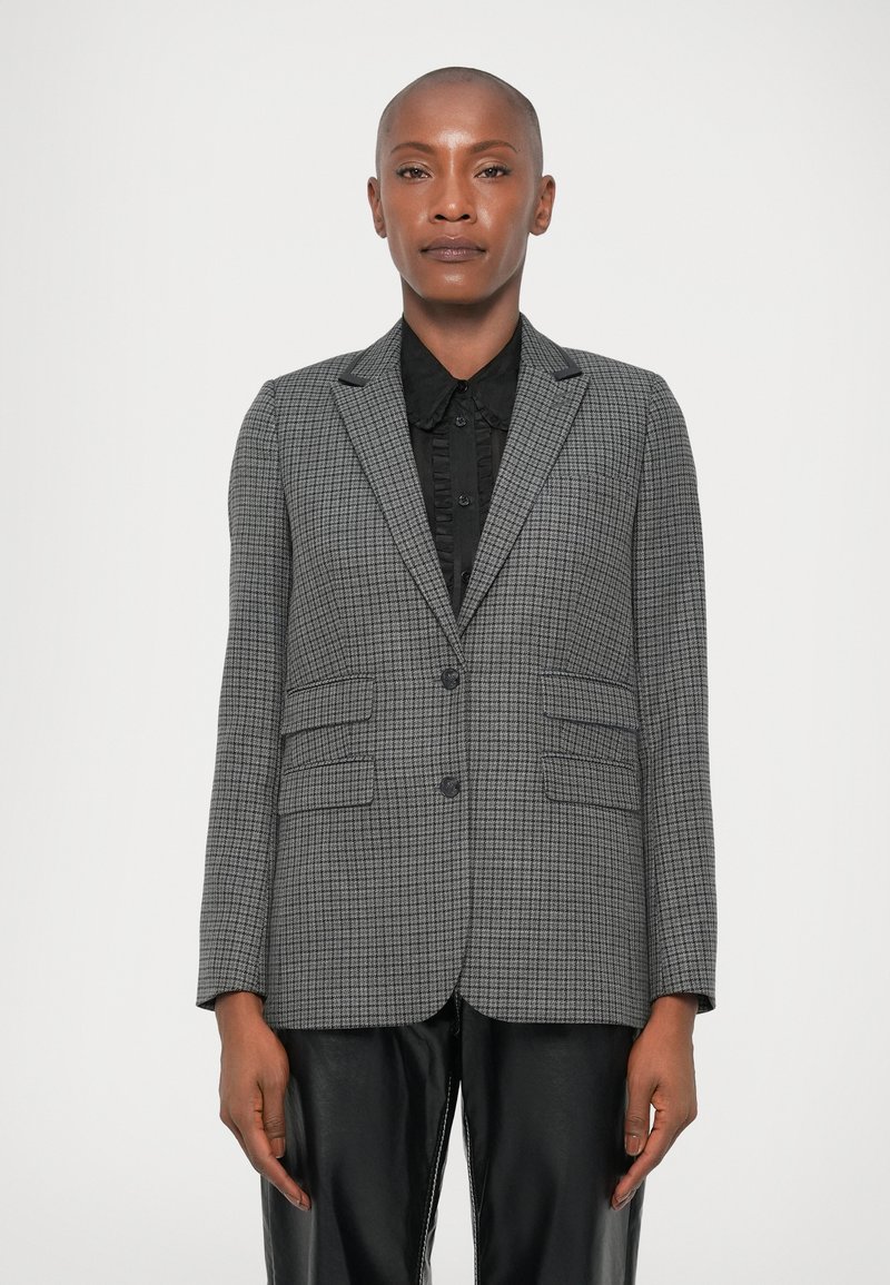 Blazer gris de cuadros con solapas de pico, dos bolsillos delanteros y cierre de un botón, usado sobre una camisa negra y pantalones de cuero.