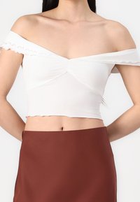 Bílý crop top bez ramínek s volánovými okraji a uzlovým designem uprostřed. Nosený s hnědou sukní s vysokým pasem.