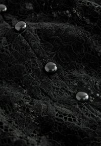 Sort blonde stof med intrikate blomsterdesigns, prydet med glatte sorte knapper til lukning langs kanten. Teksturert og lagdelt design.