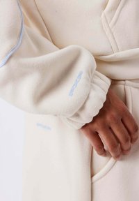 Cremefarbener Pullover mit gerippter Textur, lockeren Bündchen und blauen Akzenten. Mit gesticktem Logo am Ärmel und Kängurutasche. Weiches Material.
