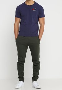 Muž v námořnickém modrém triku s krátkým rukávem, tmavě zelených joggingových kalhotách a bílých teniskách stojí s rukama v kapsách.