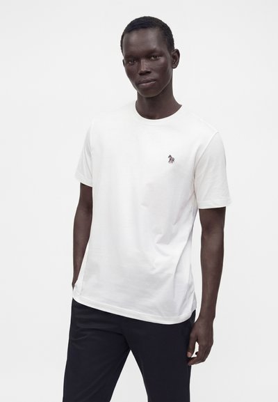 Homme portant un t-shirt blanc uni à manches courtes avec un petit logo zèbre coloré sur la poitrine et un pantalon noir, debout devant un fond blanc.