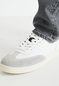 Adidași albi cu accente din suede gri, șireturi plate și detalii perforate. Purtați cu blugi din denim gri închis. Sole din cauciuc este vizibil.