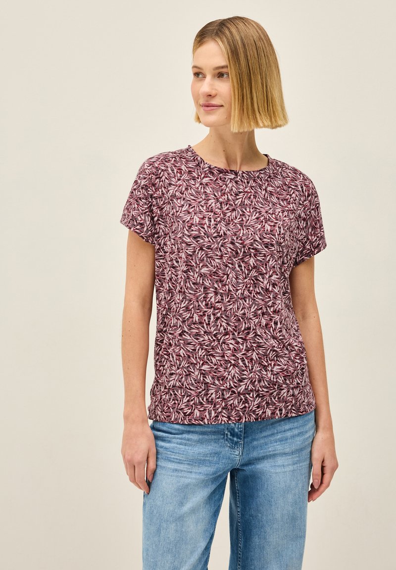 Kurzarmshirt aus burgunderfarbenem Stoff mit einem Blattmuster. Es hat einen runden Halsausschnitt und eine lockere Passform, kombiniert mit hellblauen Jeans.