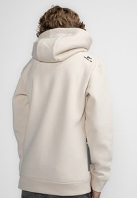Felpa beige con cappuccio realizzata in tessuto morbido, presenta un ampio cappuccio, orlo a costine e un logo sulla parte superiore della schiena.