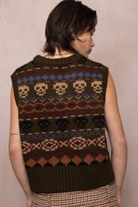 Cilvēks redzams no aizmugures, valkājot bez piedurknēm adītu veste ar daudzkrāsainām galvaskausa un ģeometriskām rakstu zīmēm uz vienkrāsaina fona.