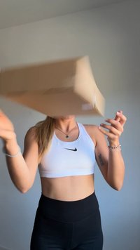 Kartona kaste, kas turēta virs personas, kas ģērbusies baltā sporta topā ar tumšu logo un melnās augstās leggingās. Fons ir gaiši pelēks.