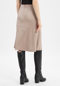 Femme portant une jupe midi beige et des bottes noires à talons haut, se tenant avec une main détendue à ses côtés, devant un fond blanc.