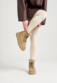 Botas altas de ante en tonalidad marrón claro con suela texturada, que cuentan con cordones negros y ojales plateados. Combinadas con leggings de color claro y una capa exterior marrón.