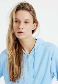 Felpa con zip di colore azzurro chiaro, realizzata in tessuto morbido. Presenta un cappuccio, un laccio e una superficie texturizzata. Design semplice con una zip frontale.