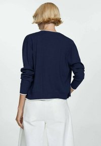 Pull en maille bleu marine à manches longues, col rond, avec des finitions blanches aux poignets. Porté avec un pantalon large de couleur claire.
