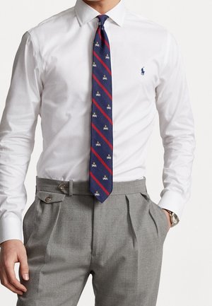 Homme portant une chemise blanche avec cravate à motifs bleu et rouge, pantalon gris plissé et montre au poignet gauche.