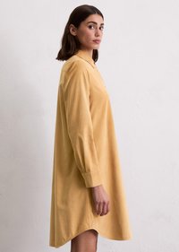 Vrouw met schouderlang bruin haar, gekleed in een lang- mouw mosterdgele jurk, staand in profiel tegen een effen witte muur.