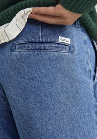 Jeans de mezclilla azul con un bolsillo trasero y una etiqueta blanca que dice "SUPERIOR JEANSWEAR". La tela parece tener una textura con un diseño clásico.