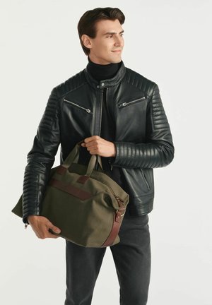 Czarna skórzana kurtka z pikowanymi ramionami, zielona torba z brązowymi skórzanymi akcentami, trzymana przez modela ubranego w ciemne dżinsy i czarną koszulę.