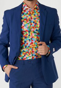 Hombre vestido con un traje azul brillante y una camisa de lunares multicolores, con una mano en el bolsillo y la otra ajustando la chaqueta.