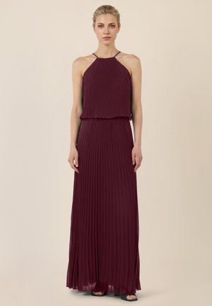 Kobieta w bezrękawną suknię maxi o ciemnoczerwonym, plisowanym materiale i wiązaniu na szyi, stojąca na tle jednolitego beżowego tła.
