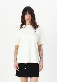 Cream oversized t-shirt made of cotton, featuring short sleeves and a graphic print on the left chest. Paired with a black mini skirt.
