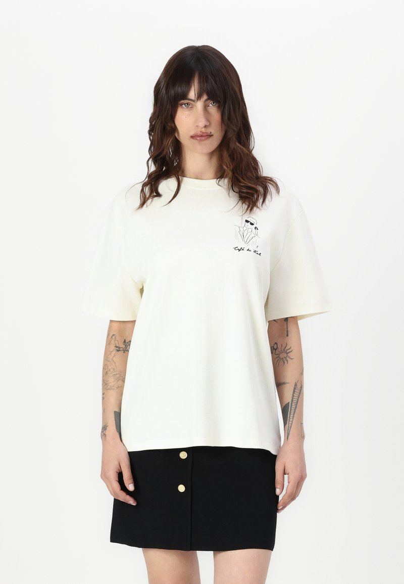 Cream oversized t-shirt made of cotton, featuring short sleeves and a graphic print on the left chest. Paired with a black mini skirt.