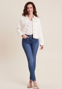 Witte denimjack met knoopsluiting, twee borstzakken; gedragen over een lichte top en donkere skinny jeans; geaccessoireerd met gouden sandalen.