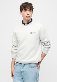 Νεαρός με κατσαρά μαλλιά που φορά ανοιχτό γκρι φούτερ Converse πάνω από μπλε πουκάμισο και ανοιχτ ό μπλε τζιν, στέκεται με το ένα χέρι στην τσέπη.