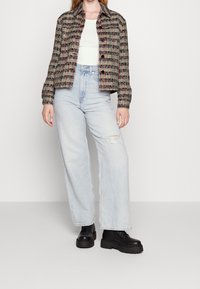 Woven jacket in multicolored fibers, featuring a collared neckline, button closure, and ribbed sleeves. Paired with light blue wide-leg jeans.