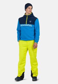 Sininen fleece-pusero, jossa tumman laivastonsininen yläosa, kirkas keltainen vetoketju ja "ROSSIGNOL" -logo. Yhdistetty kirkkaan keltaisten housujen kanssa ja mustilla yksityiskohdilla.