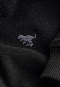 Tissu noir au toucher lisse avec un petit motif de dinosaure gris brodé. Détails de poignet côtelé ajoutent de la dimension.
