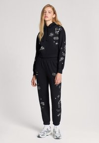 Completo felpa nera con pantaloni jogger, presenta grafiche bianche, cappuccio con cordino, polsini a coste e cintura elastica.