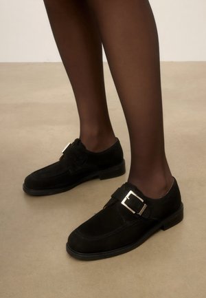 Jambes en collants sombres transparents portant des chaussures monk en daim noir avec des boucles argentées sur un sol beige.