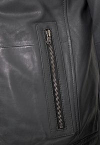 Schwarze Lederjacke mit einer Reißverschlusstasche, strukturiertem Oberflächenlayout und genähten Akzenten. Der Reißverschluss ist metallisch und befindet sich an der Vorderseite.