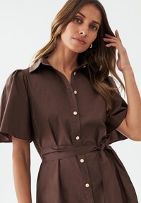 Abito camicia marrone con maniche corte a sbuffo, chiusura frontale con bottoni e vita legata. Realizzato in un tessuto morbido e leggero.