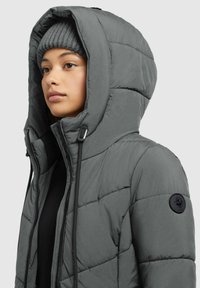Doudoune grise avec une grande capuche, texture matelassée, accents noirs et un patch logo sur la manche. La personne porte un bonnet en tricot gris.