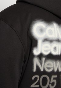 Tessuto nero con testo sprayato bianco e grigio che legge "Calv Jeans New 205" su un capo piegato, mostrando una cucitura della spalla.