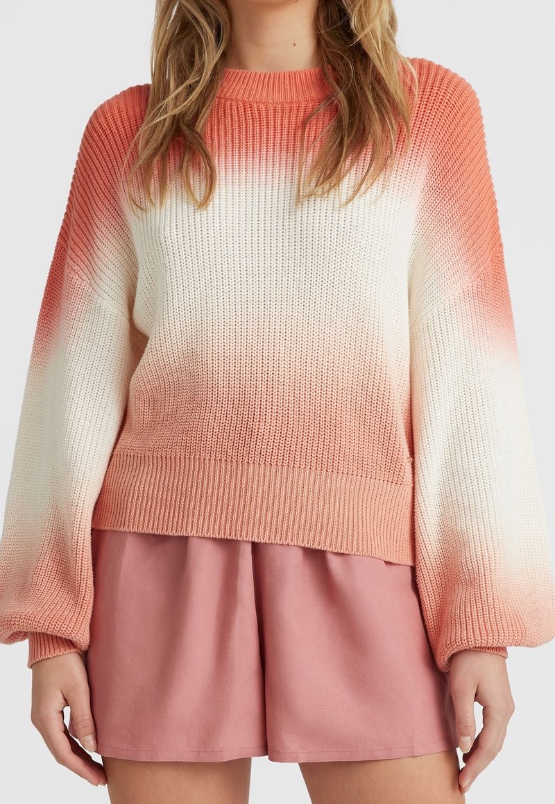 Femme portant un pull tricoté dégradé rose et blanc avec de longues manches bouffantes et un short rose ample sur un fond uni.