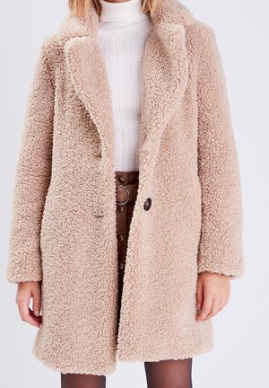 Femme portant un manteau teddy texturé beige sur un pull col roulé blanc et une jupe marron taille haute boutonnée.
