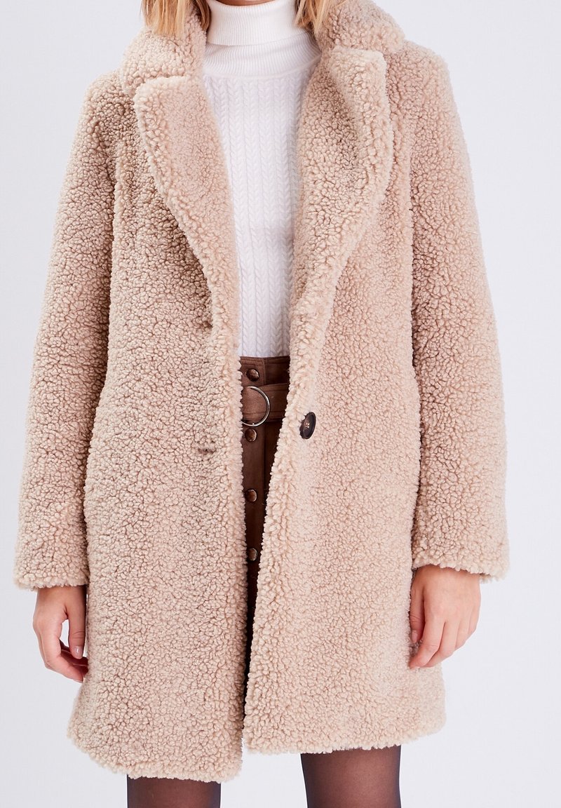 Femme portant un manteau teddy texturé beige sur un pull col roulé blanc et une jupe marron taille haute boutonnée.