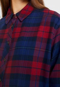 Rutig skjorta med knappar i röd, marinfärgad och grön färg. Mjuk tyg med pekad krage och kontrasterande knappar, som visar ett tartanmönster.