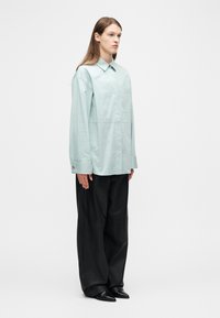 Mujer de pie con una camisa grande de color menta claro, pantalones anchos negros y zapatos negros puntiagudos, sobre un fondo blanco liso.
