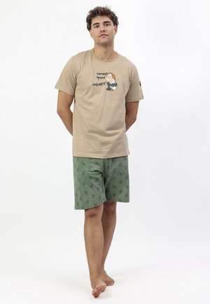 Joven de pie descalzo con camiseta beige con dibujo animado y texto, vistiendo pantalones cortos verdes con pequeños patrones impresos sobre un fondo blanco.